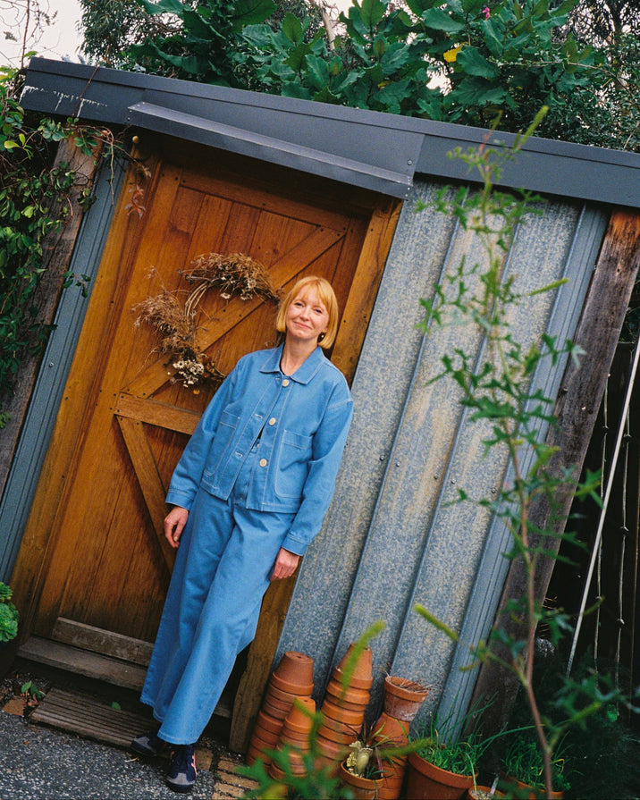 A person wearing a matching blue denim jacket and wide-leg pants set, standing outdoors next to a wooden door and stacked terracotta pots.