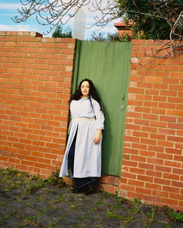 A woman stands in front of a red brick wall and a green wooden door, wearing a long white and blue striped dress with a beige belt. Bare tree branches hang from above, and the ground is paved with cobblestones.