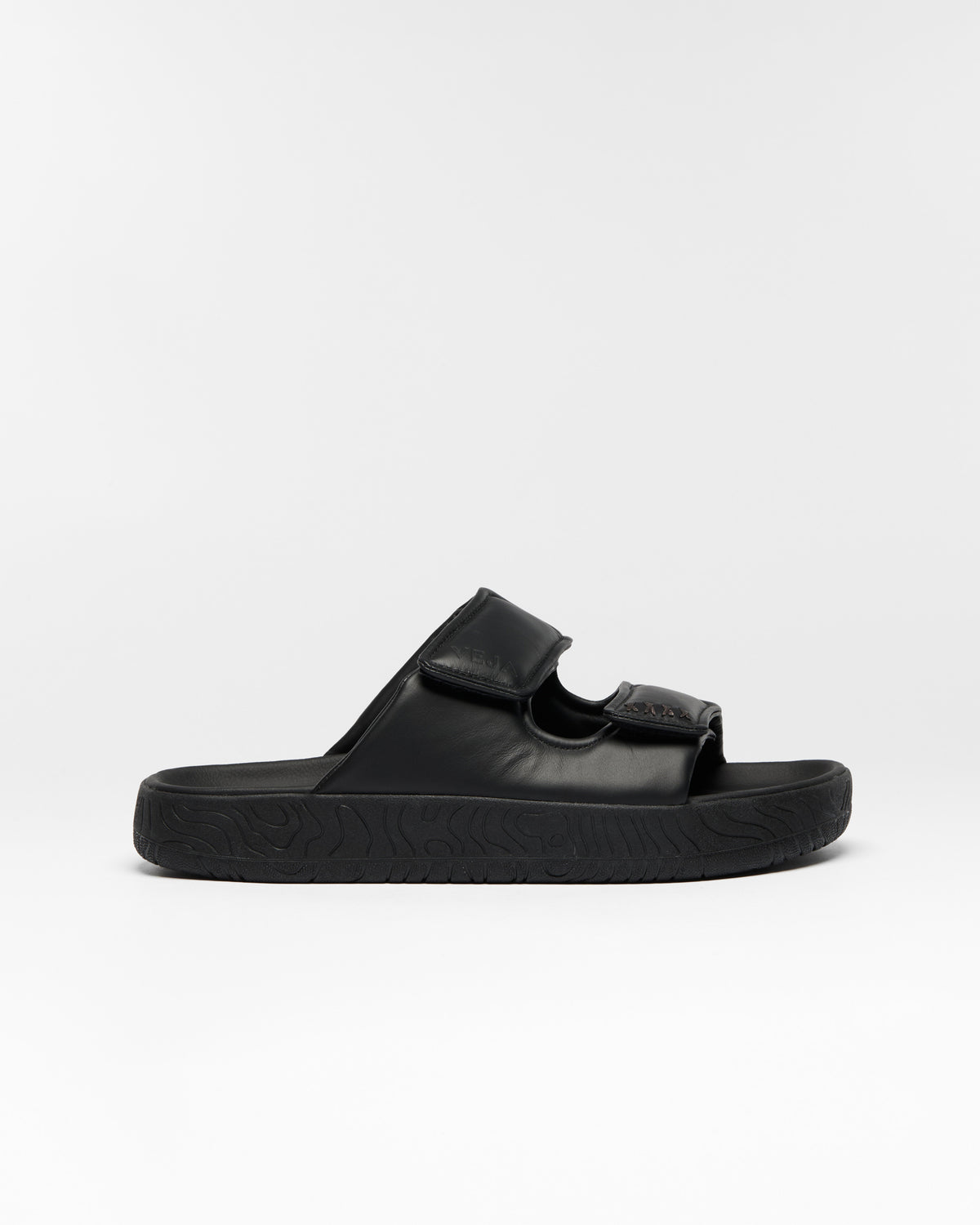 A single black slide sandal with a wide strap and a textured sole, shown in profile against a light gray background.