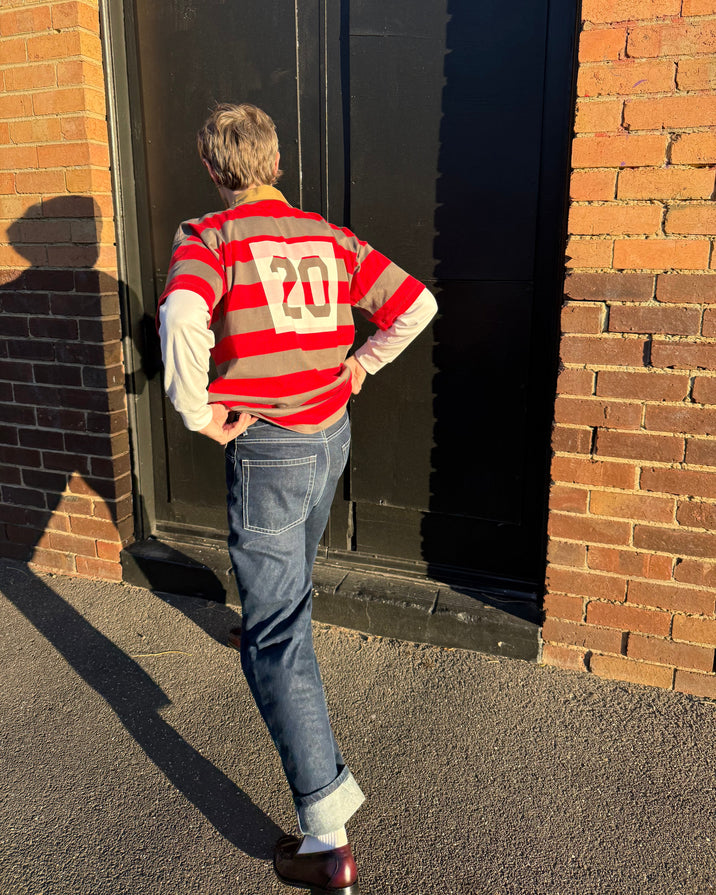 A person standing outdoors in front of a brick building and dark door, wearing a red and beige striped long-sleeve sweater with a large white number 20 on the back, blue jeans, and brown shoes.