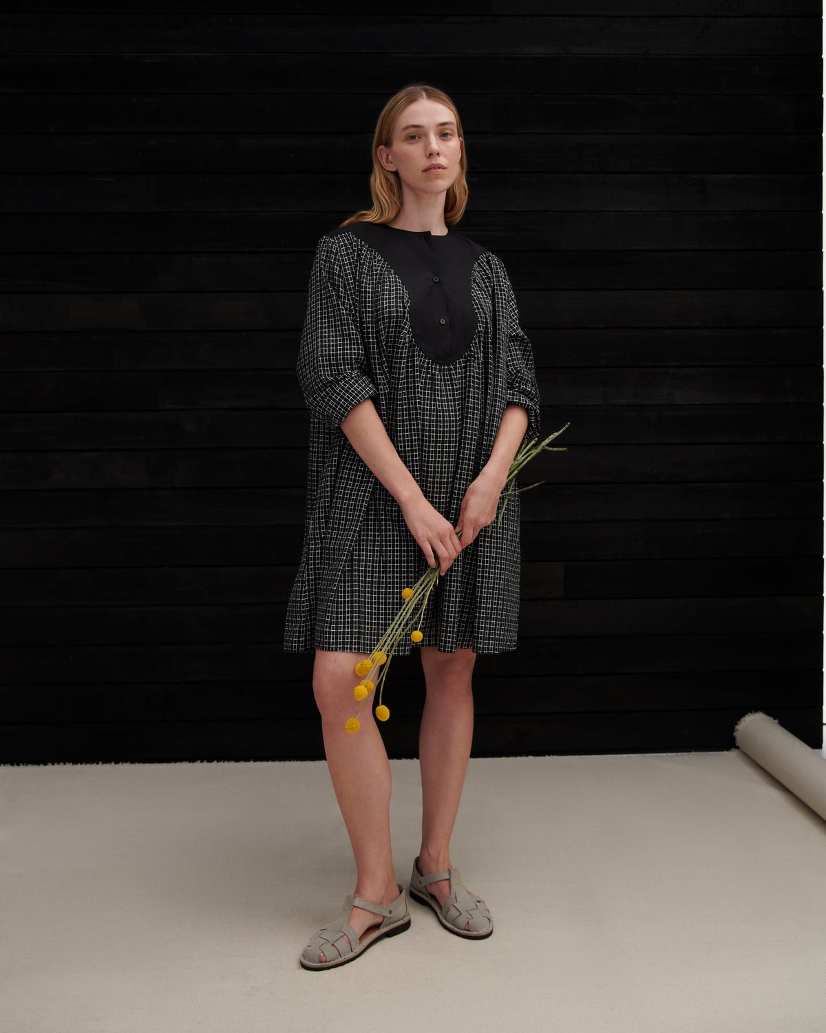 A woman stands against a black wood plank wall wearing a black and gray gingham tunic dress with a solid black yoke and holding a small sprig of yellow flowers.