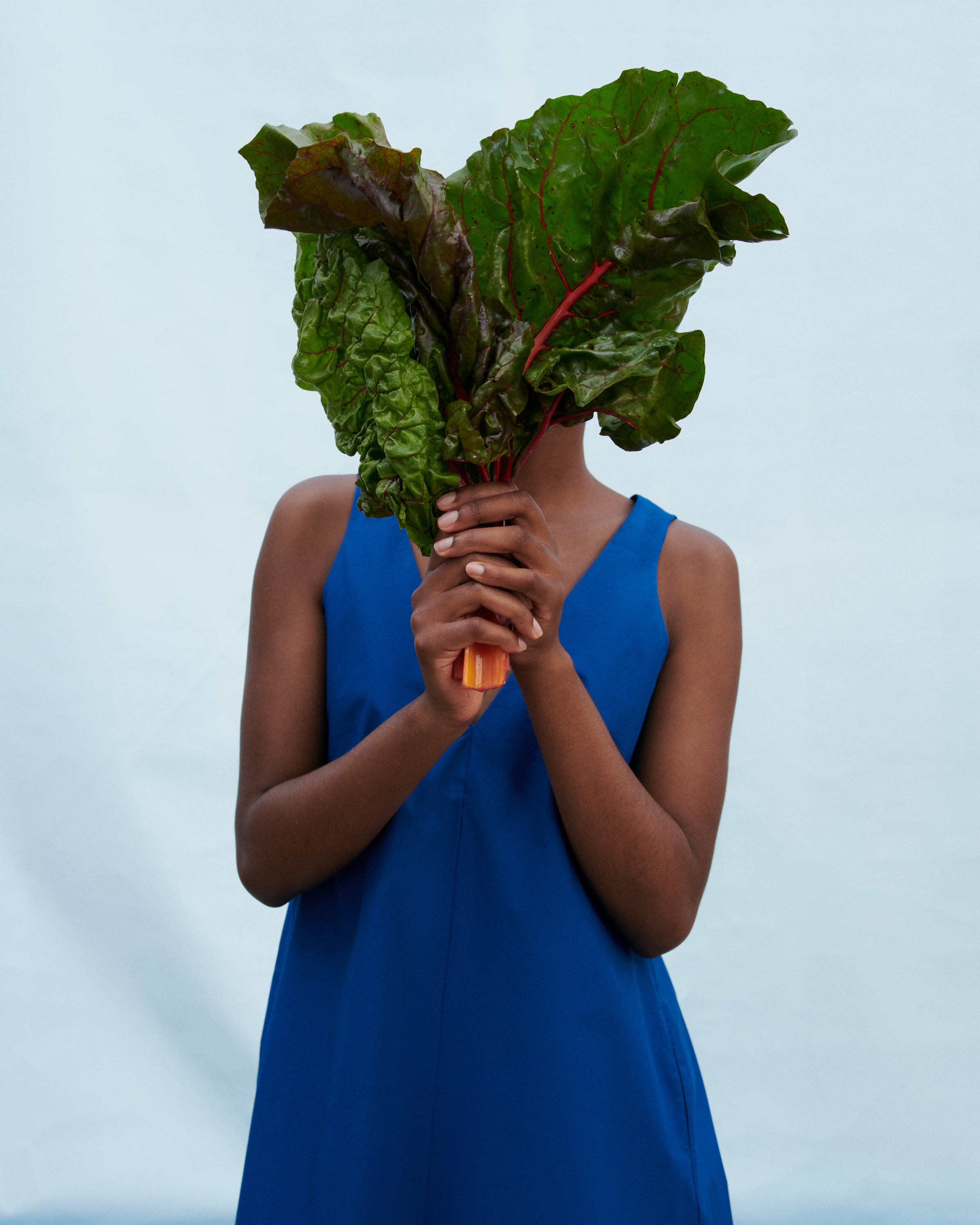 A person in a bright blue sleeveless dress holds a large bunch of fresh green rhubarb in front of their face against a pale blue studio background.