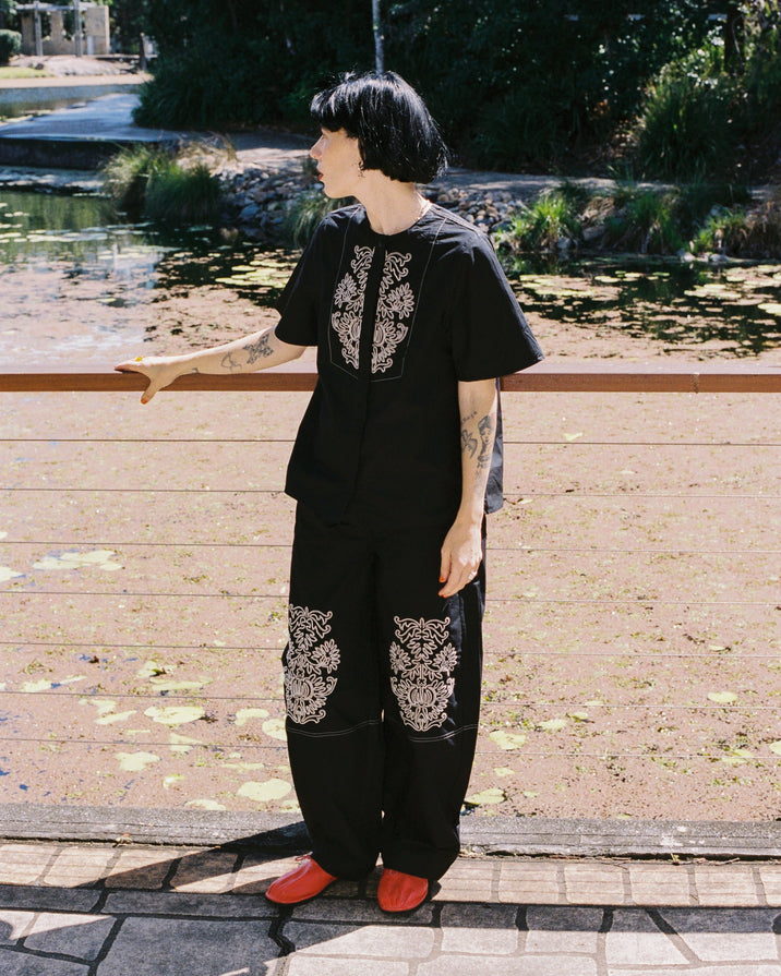 A person standing outdoors near a pond wearing a black short-sleeve top and matching loose pants with ornate white embroidered or printed patterns on the chest and upper legs, paired with bright red shoes.