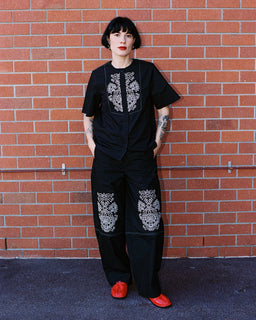 A person with short black hair and red lipstick stands against a red brick wall wearing a black short-sleeve top and matching wide-leg pants with white floral embroidery, paired with bright red shoes.