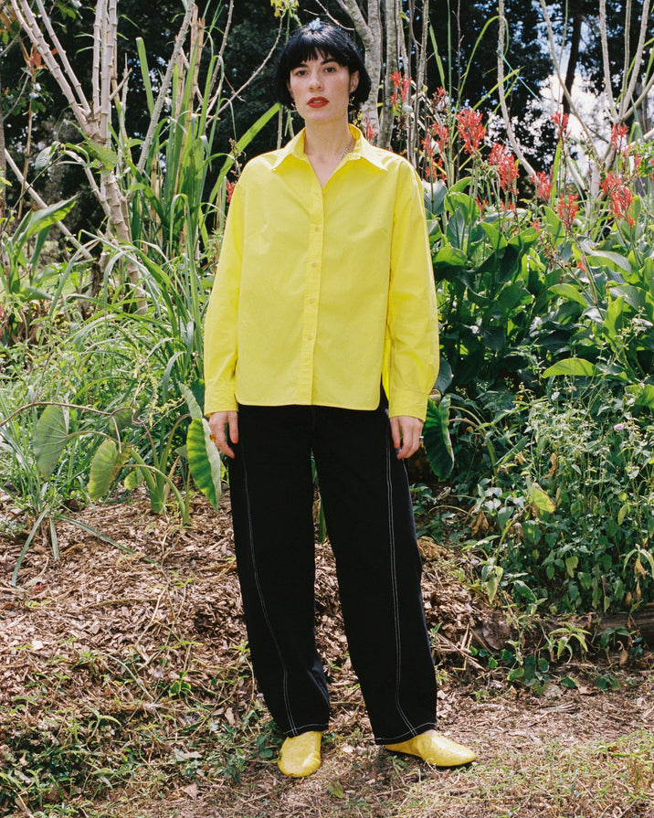 A person standing outdoors in a garden wearing a bright yellow button-up shirt, black wide-leg pants, and matching yellow shoes.