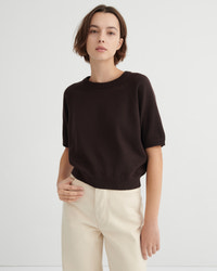 A model wearing a dark brown short-sleeve crewneck sweater and cream-colored trousers, standing against a light gray studio background.