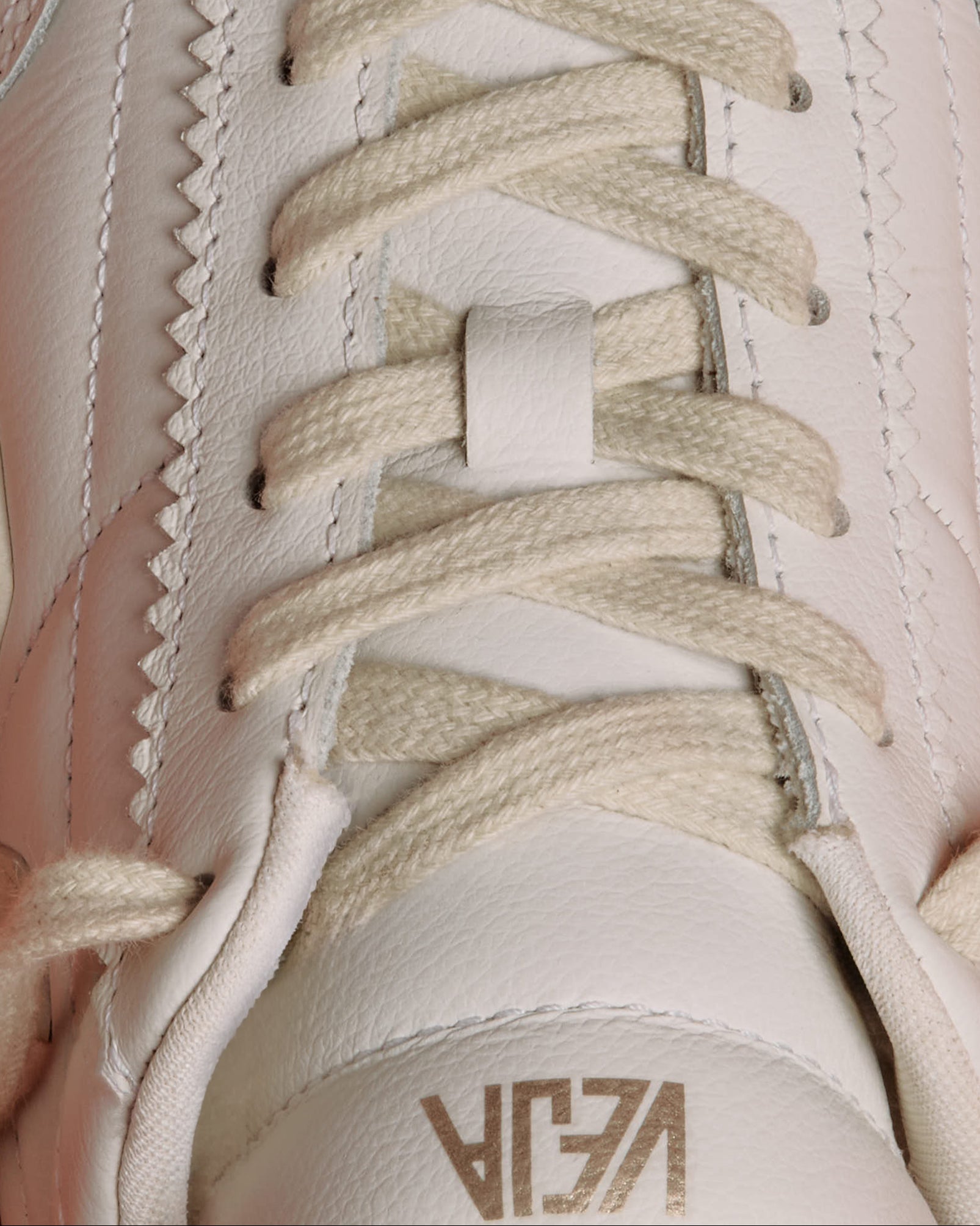 Close-up of a white leather sneaker with cream-colored laces and a metallic gold embossed Veja logo on the tongue.