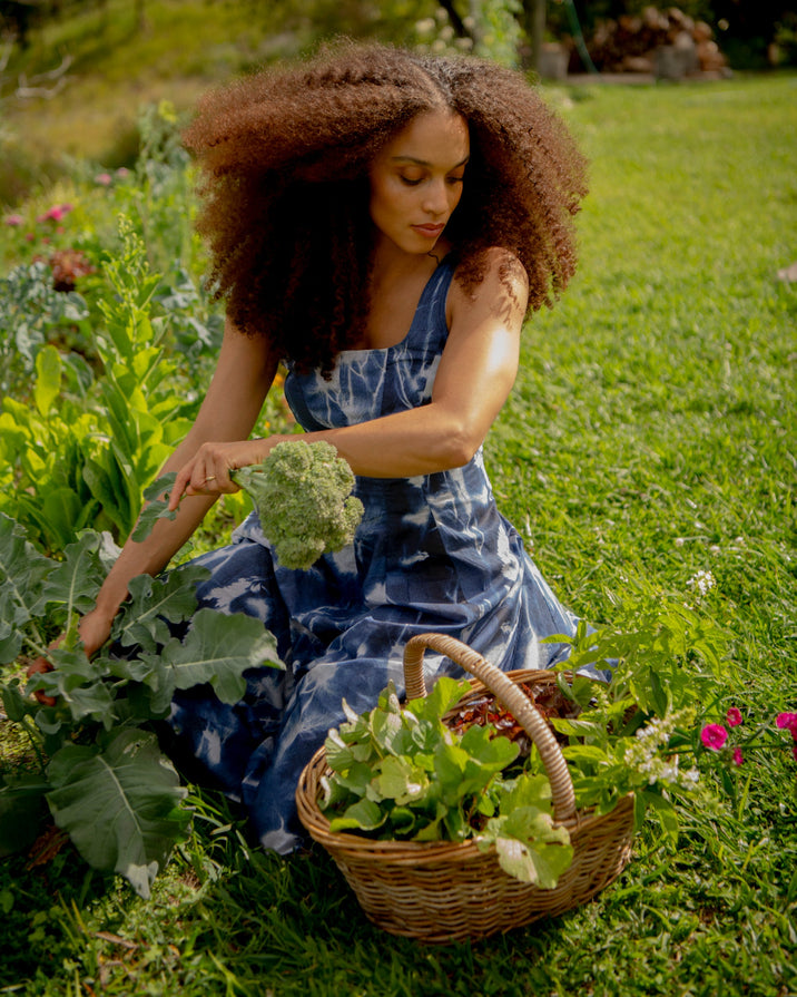 A woman with voluminous curly hair sits in a grassy garden, harvesting green broccoli from a plant while wearing a blue and white patterned dress. A wicker basket filled with leafy greens sits beside her.