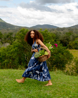 A woman with long curly hair walks barefoot across a grassy field, wearing a blue and white tie-dye maxi dress and carrying a wicker basket filled with green vegetables and pink flowers, with mountains and a cloudy sky in the background.