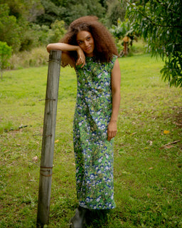 A woman with curly brown hair stands outdoors in a grassy field leaning against a wooden post, wearing a sleeveless, knee-length green dress with a blue and purple floral pattern.