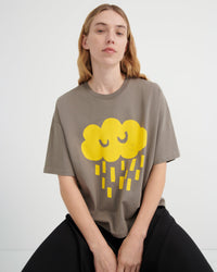 A woman wearing an oversized taupe-gray t-shirt with a yellow cartoon cloud graphic, paired with black pants, seated against a light gray studio background.