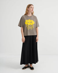 A woman modeling a taupe short-sleeve t-shirt with a yellow cloud and rain graphic, paired with a long black skirt and black shoes, against a plain white studio background.