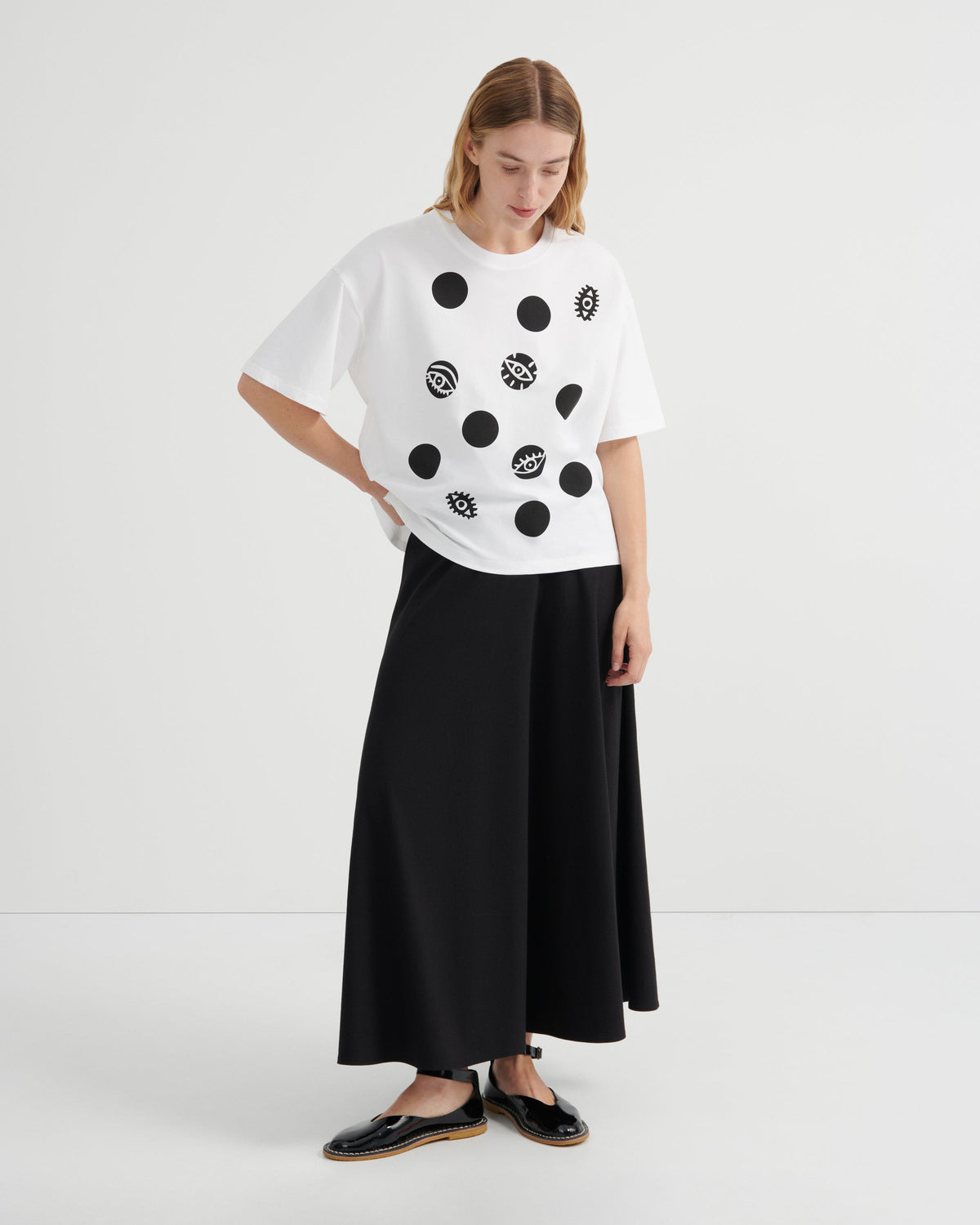 A model stands against a light gray studio background wearing a white oversized T-shirt with black circular dot and pattern graphics, a long black skirt, and black flat shoes.