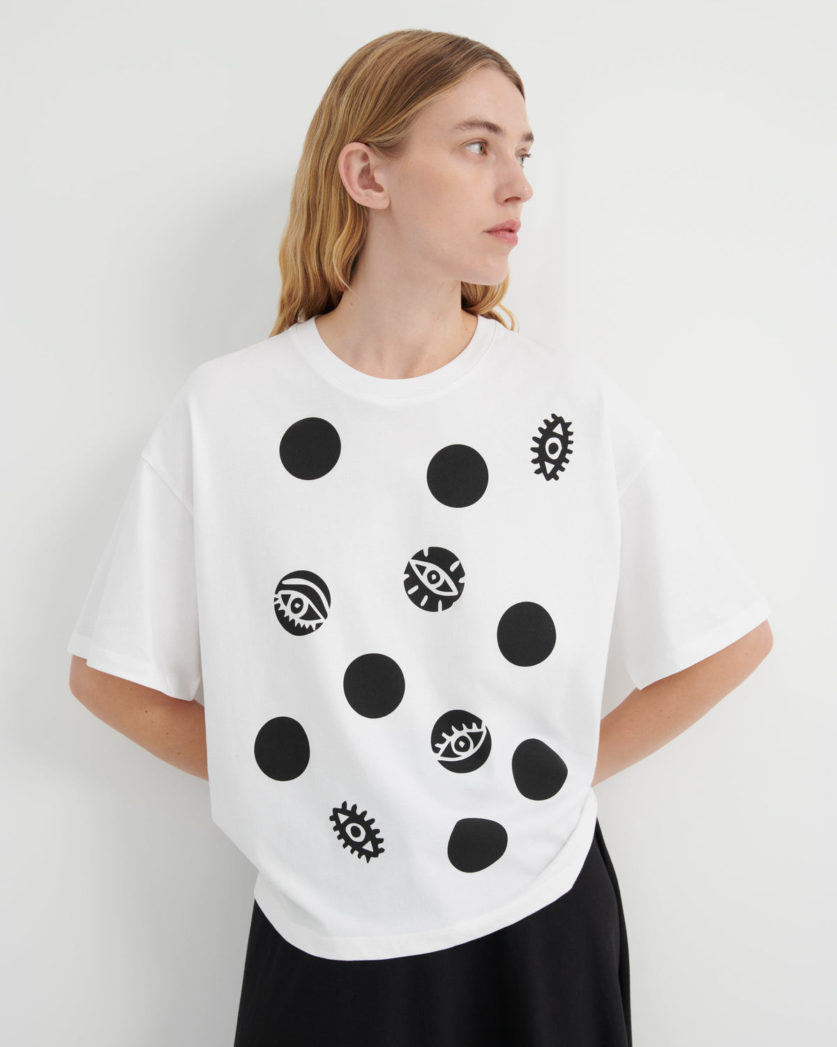 A woman wearing a white oversized T-shirt with black polka dots and eye-and-gear motifs, paired with black bottoms, photographed against a plain light gray studio background.