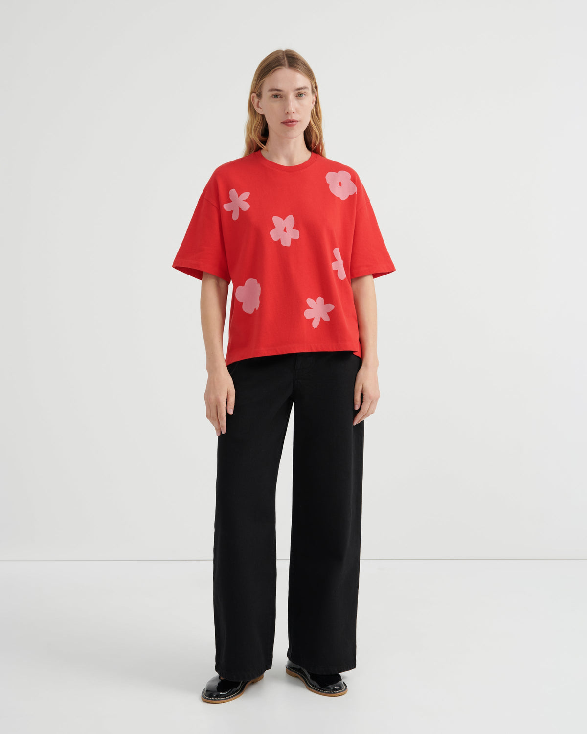 A woman stands against a plain light gray studio background wearing a bright red short-sleeve T-shirt with large pale pink flower motifs and black wide-leg pants.