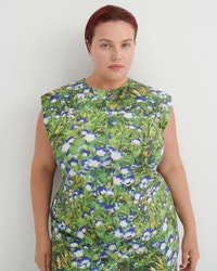 A woman with short auburn hair wearing a sleeveless, high-neck dress with a green, blue, and white floral pattern, standing against a plain light gray background.
