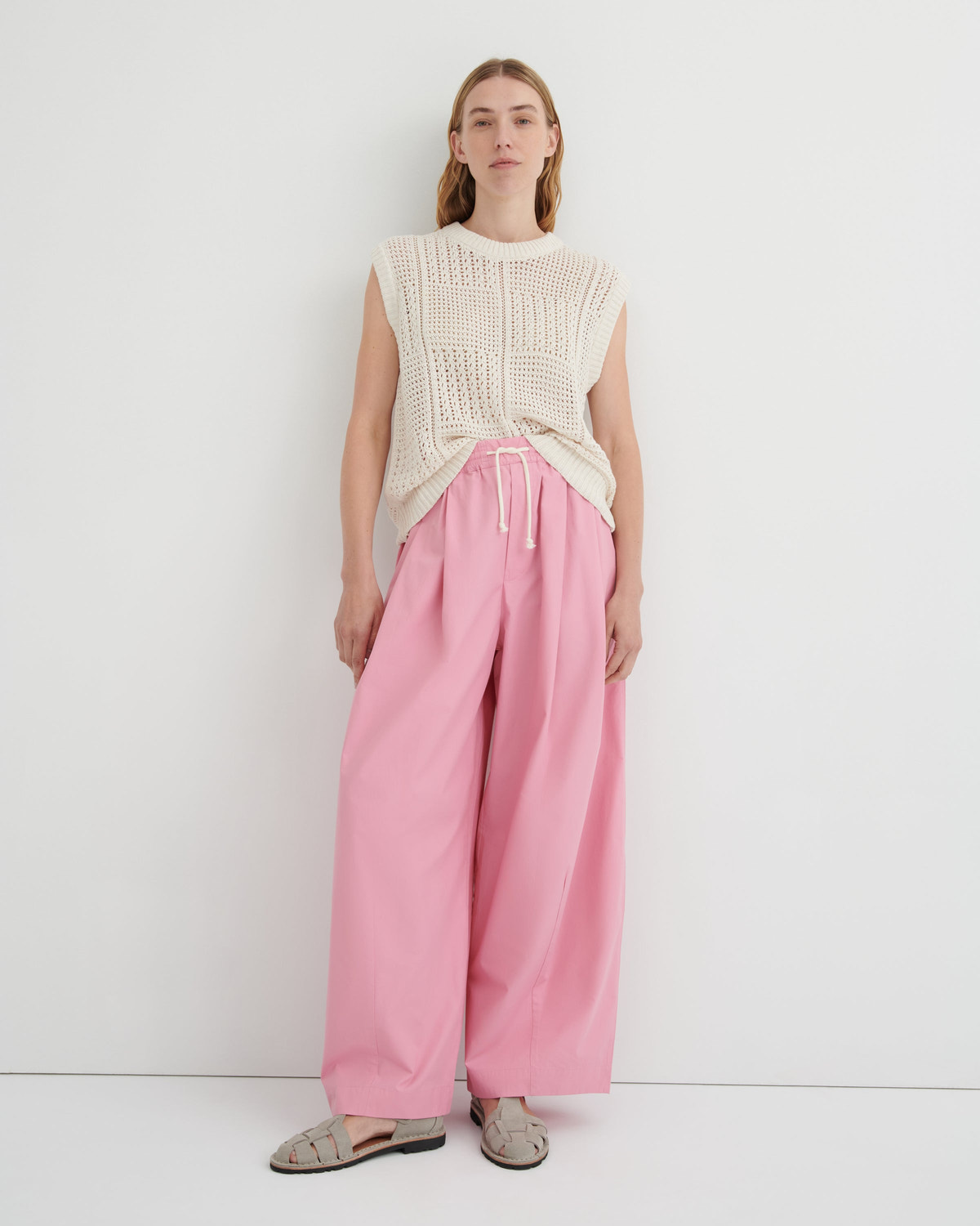 A model stands against a plain white studio background wearing a cream sleeveless knit top, pink wide-leg drawstring pants, and beige flat sandals.