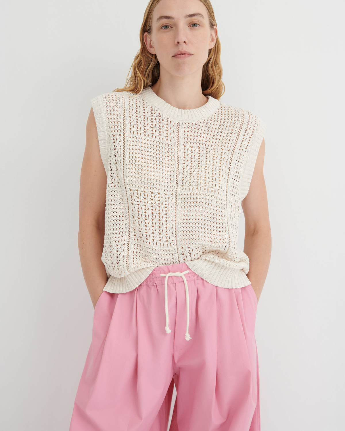 A model wearing a cream-colored sleeveless open-knit sweater vest paired with pink drawstring trousers, standing against a plain light gray studio background.