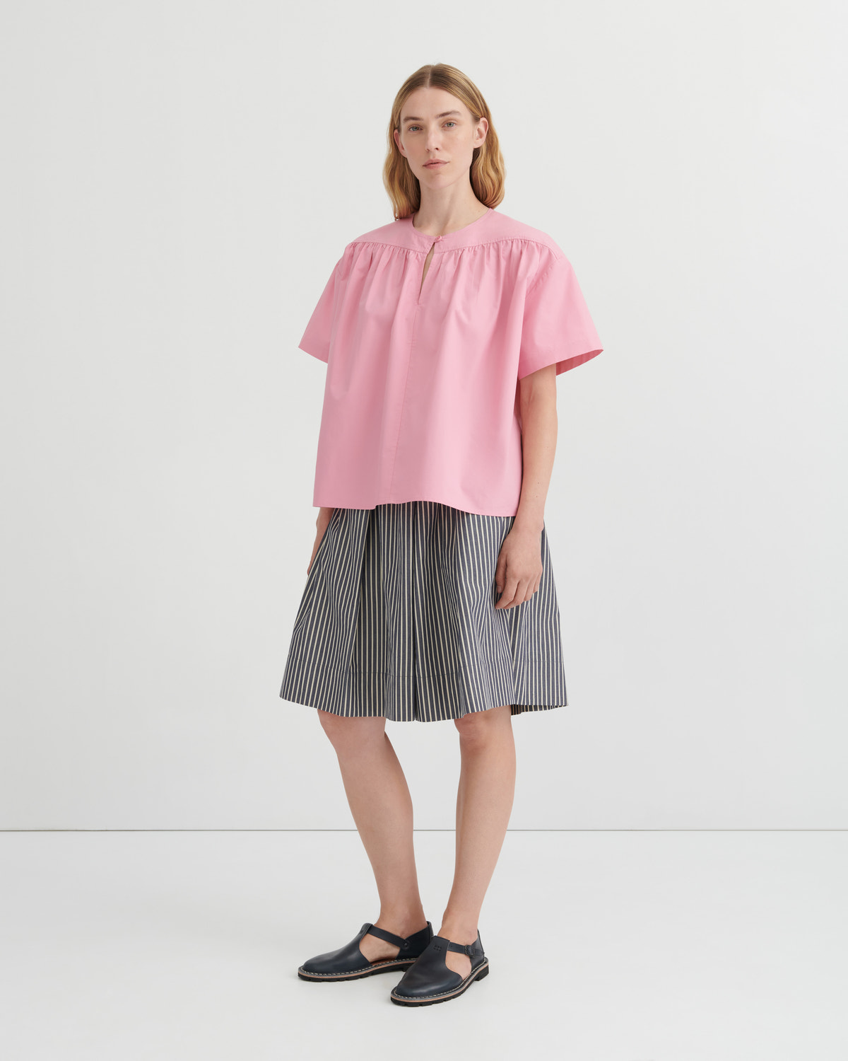 A woman modeling a light pink short-sleeve blouse with a gathered neckline and pleated skirt in a studio setting.