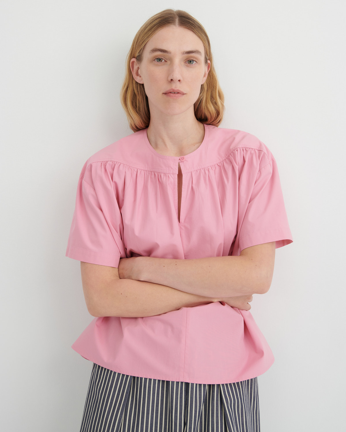 A woman wearing a light pink short-sleeve blouse with a gathered round neckline and a split V-front detail, paired with black and white striped trousers, standing against a light gray wall.
