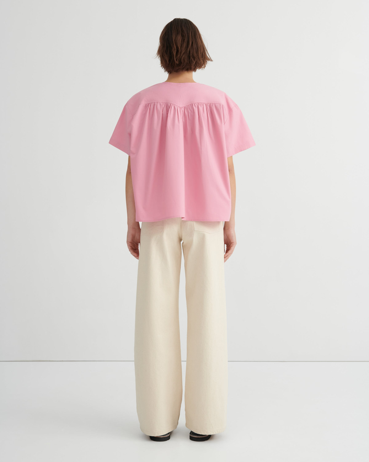 A woman stands backwards wearing a light pink short-sleeve blouse with a gathered neckline and wide-leg cream trousers, paired with red sandals, against a clean white studio background.