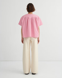 A woman stands backwards wearing a light pink short-sleeve blouse with a gathered neckline and wide-leg cream trousers, paired with red sandals, against a clean white studio background.