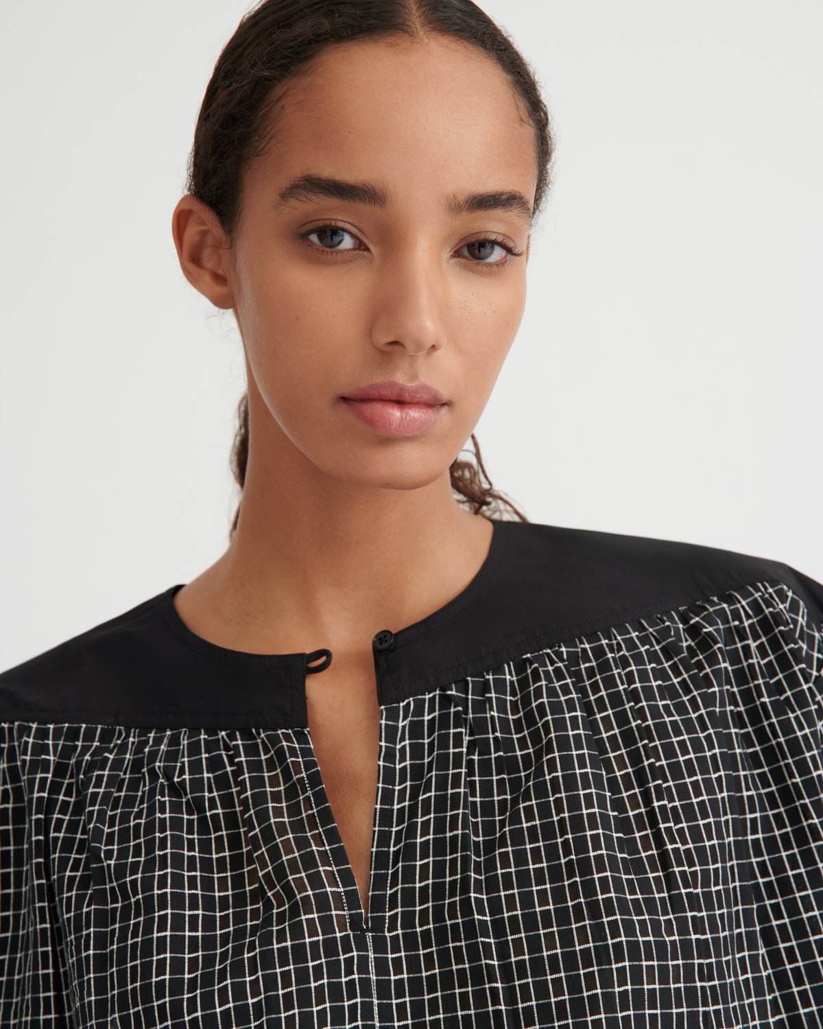 A close-up of a model wearing a black and white checkered top with a solid black collar and button placket, set against a plain light gray background.
