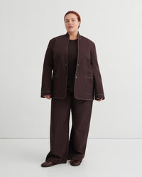 A model stands against a light gray studio background wearing a dark brown matching set consisting of a cropped jacket with contrast light piping, wide-leg trousers, and a black top.