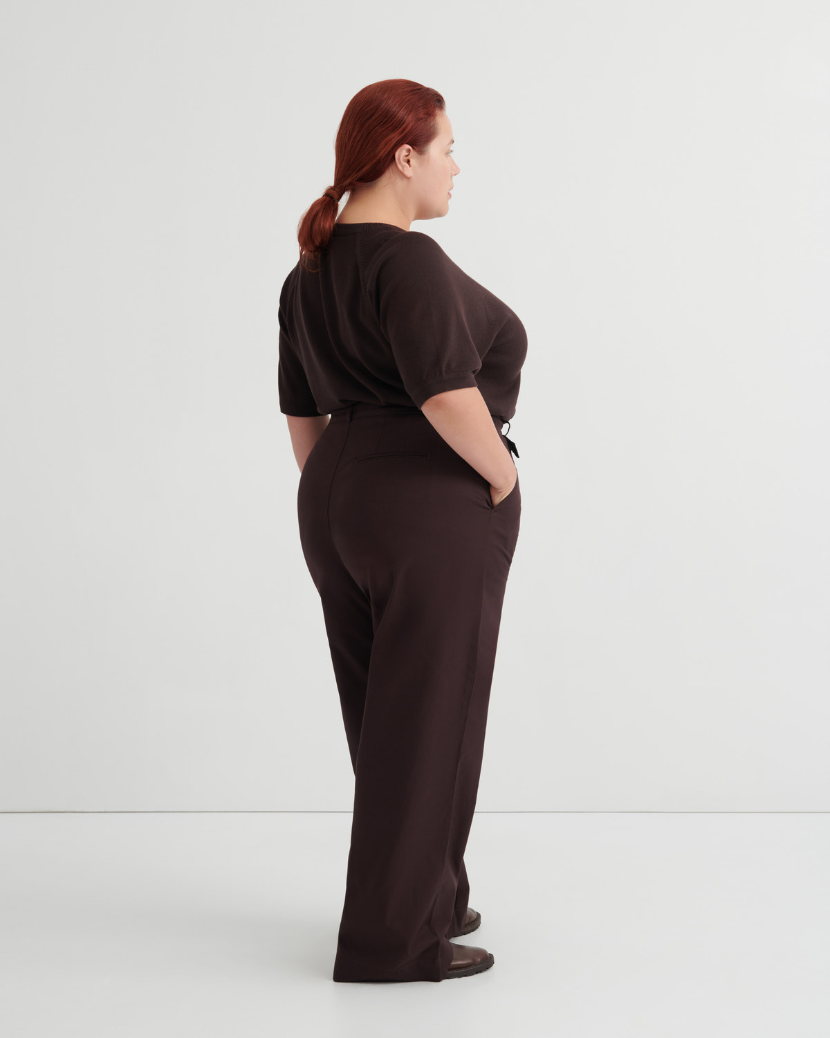 A woman with red hair tied back is shown from the back wearing a dark brown short-sleeve top and matching dark brown wide-leg trousers, standing against a plain light gray studio background.