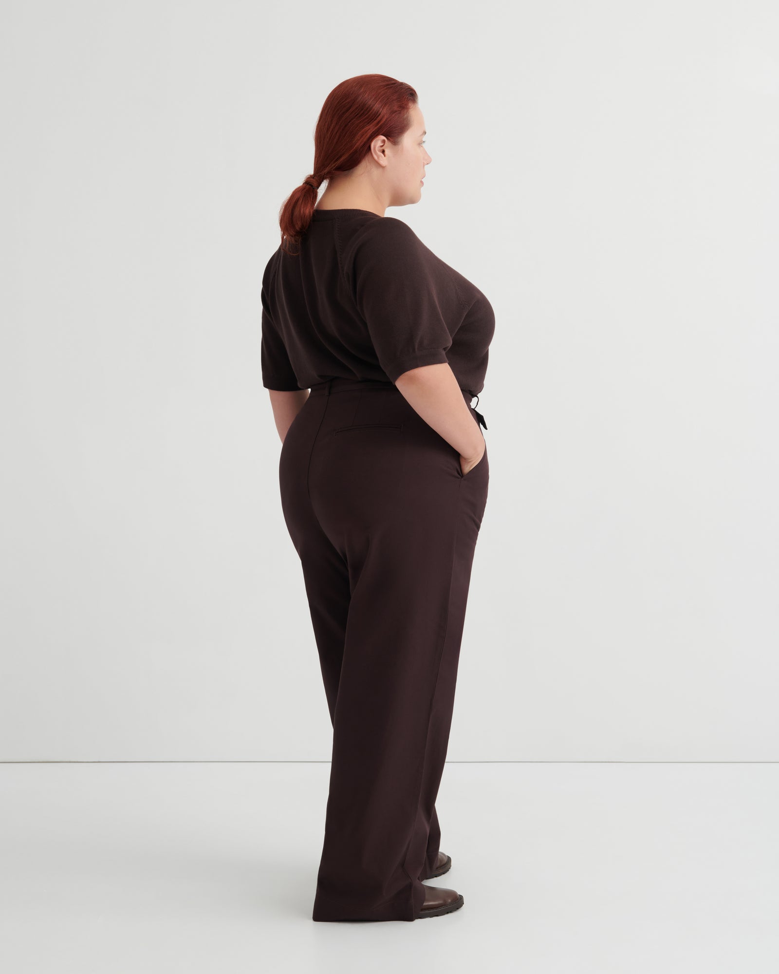 A woman with red hair tied back is shown from the back wearing a dark brown short-sleeve top and matching dark brown wide-leg trousers, standing against a plain light gray studio background.
