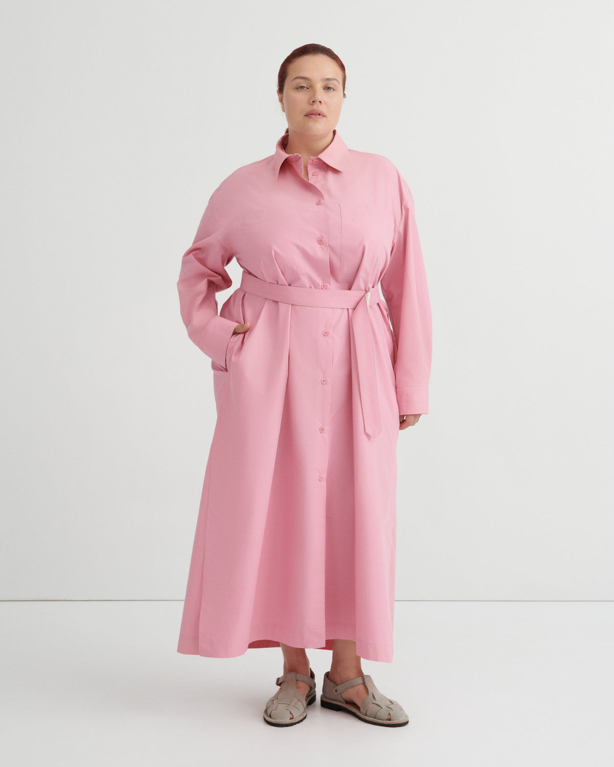 A woman modeling a pink long-sleeve shirt dress with a matching waist tie belt, standing against a plain white studio background.