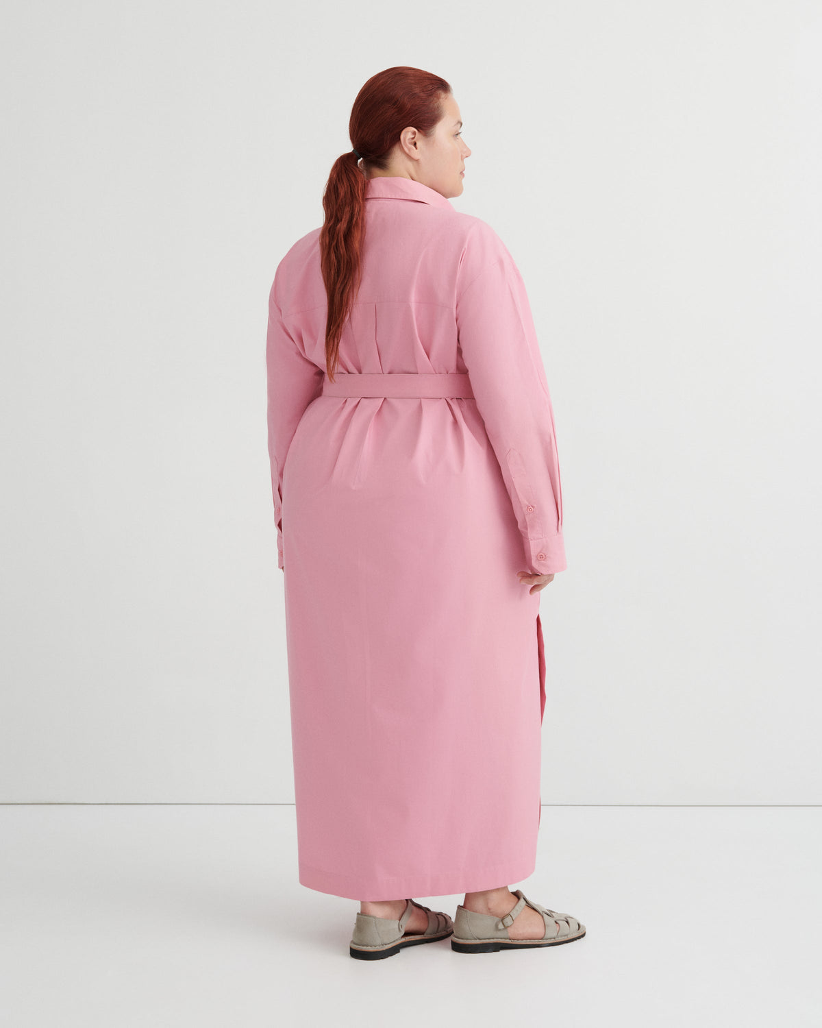 A woman with red hair tied back in a ponytail is shown from the back wearing a long pink belted coat or dress over a light gray top and gray sandals, photographed against a clean white studio background.