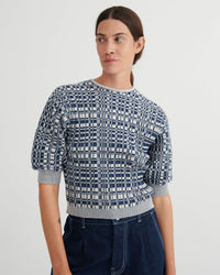 A woman wearing a short-sleeve knit top with a navy and white geometric grid pattern, paired with dark blue jeans, against a plain light gray studio background.