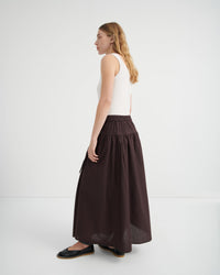 A woman shown from the side profile wearing a white sleeveless top and a long, dark brown gathered skirt.