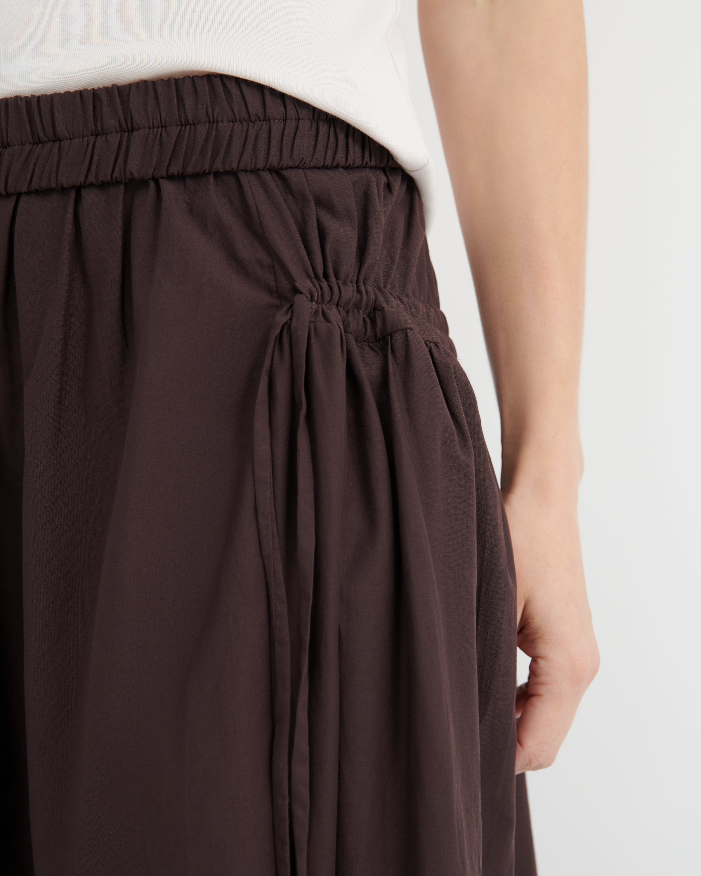 Close-up of a person wearing dark brown pleated skirt with an elastic waistband, paired with a white top against a light gray background.