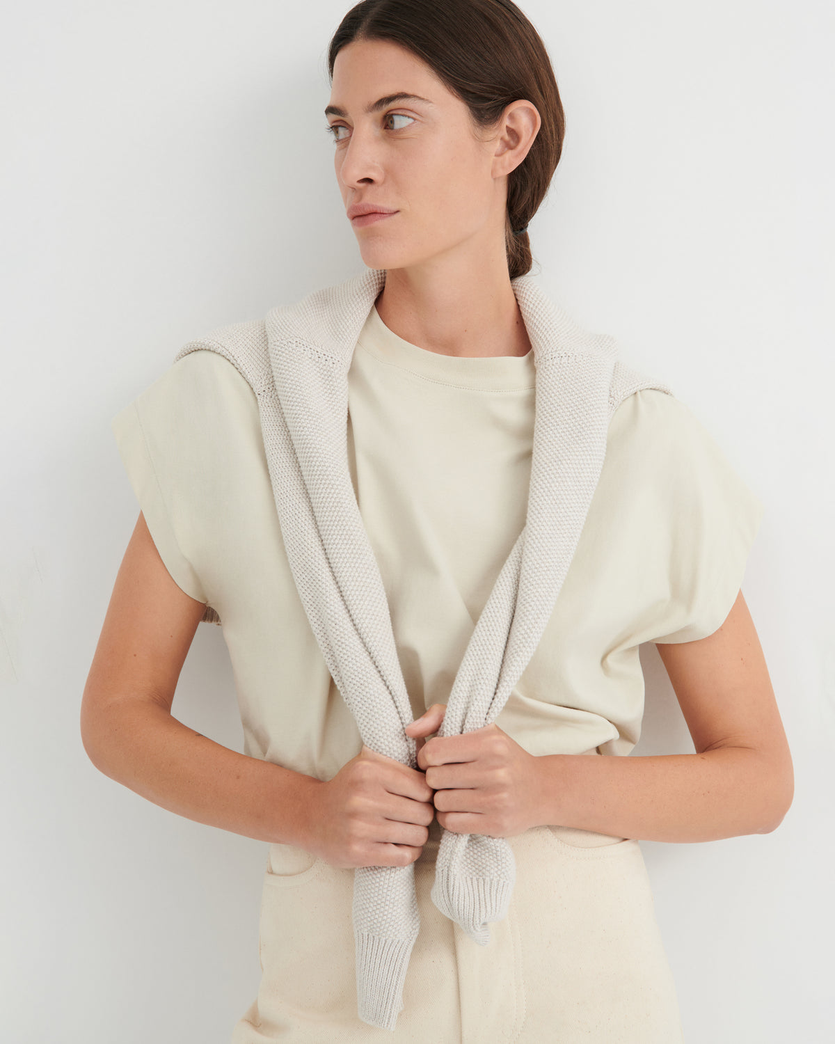 A woman wearing a cream-colored knit cardigan with a shawl collar over a matching cream t-shirt and trousers, standing against a plain white wall.