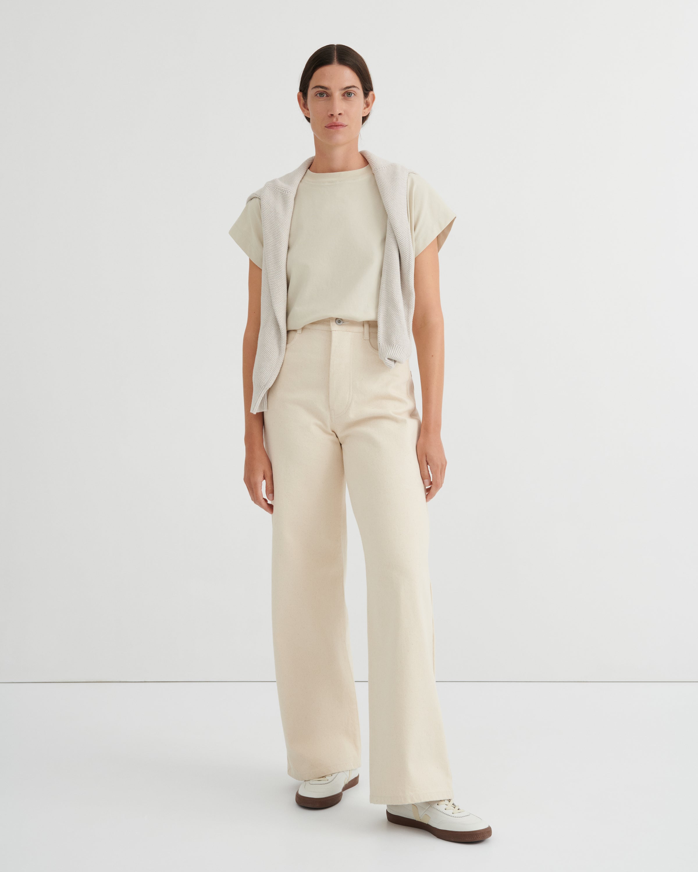 A model stands against a clean white studio background wearing a light beige short-sleeve top, matching wide-leg trousers, a draped light gray cardigan, and white sneakers.
