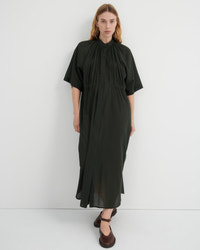 A woman modeling a dark olive green midi dress with a high gathered neckline, short puff sleeves, and a button-front bodice, standing against a plain light gray studio background.