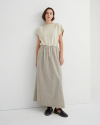 A woman stands against a plain white wall wearing a cream short-sleeve top and a beige-and-white vertically striped maxi skirt with a drawstring waist, paired with black slip-on shoes.