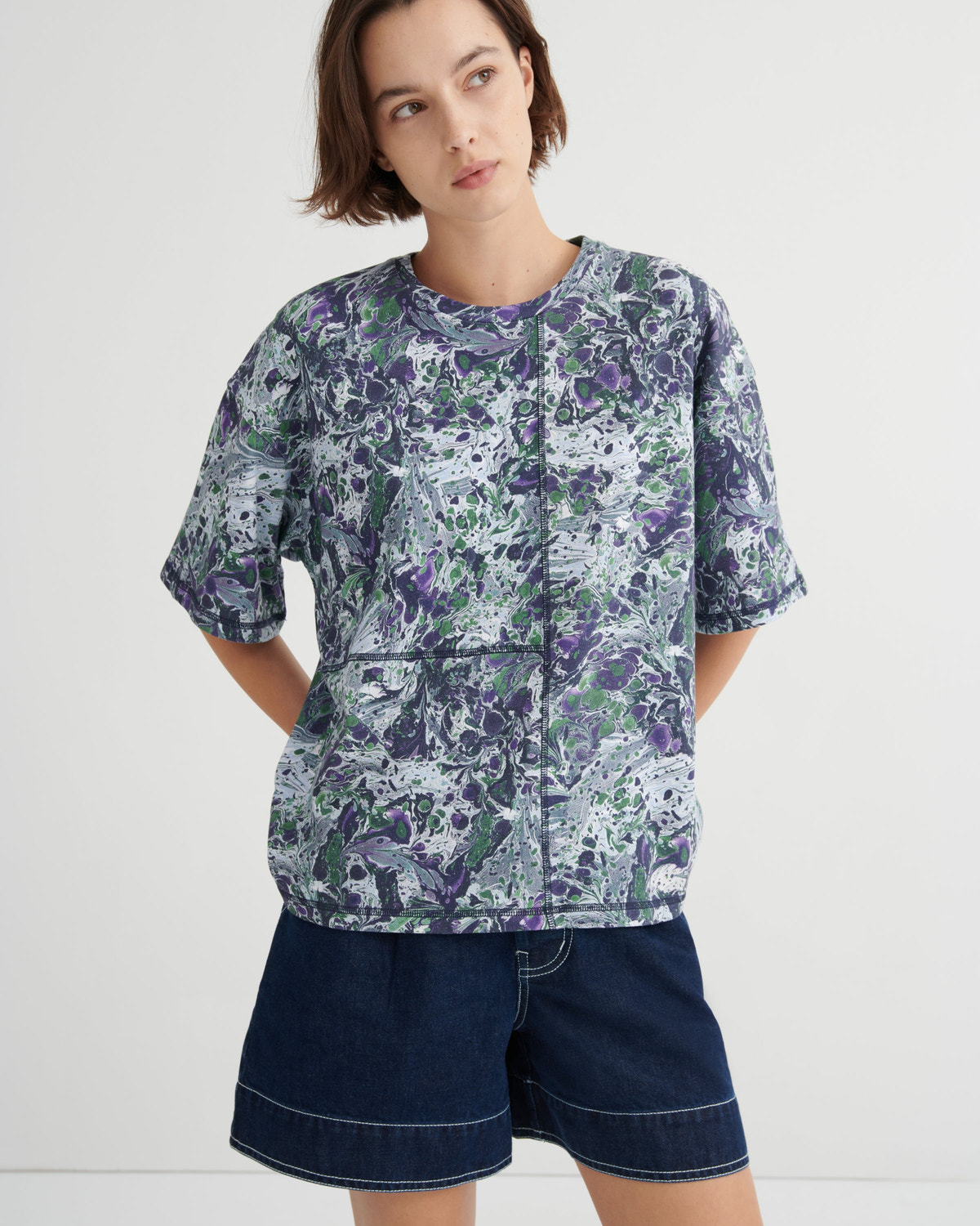 A woman modeling a short-sleeve button-front top with an abstract marbeld print in purple, green, and white, paired with dark denim shorts against a plain light gray studio background.