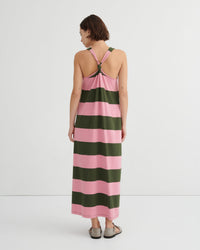 A woman viewed from the back wearing a sleeveless maxi dress with wide horizontal pink and dark green stripes and a crossover halter-style back, against a clean white studio background.