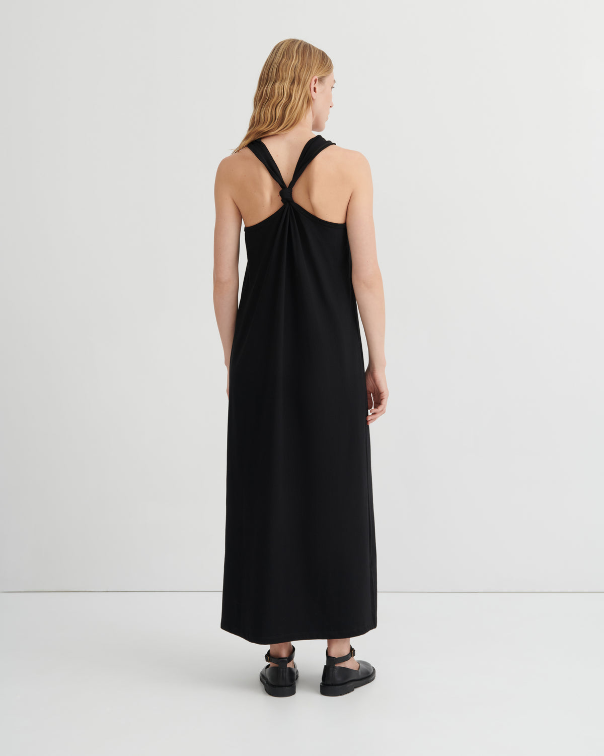 A woman viewed from the back wearing a sleeveless black maxi dress with a crossover back design, paired with black flat shoes, against a plain light gray studio background.