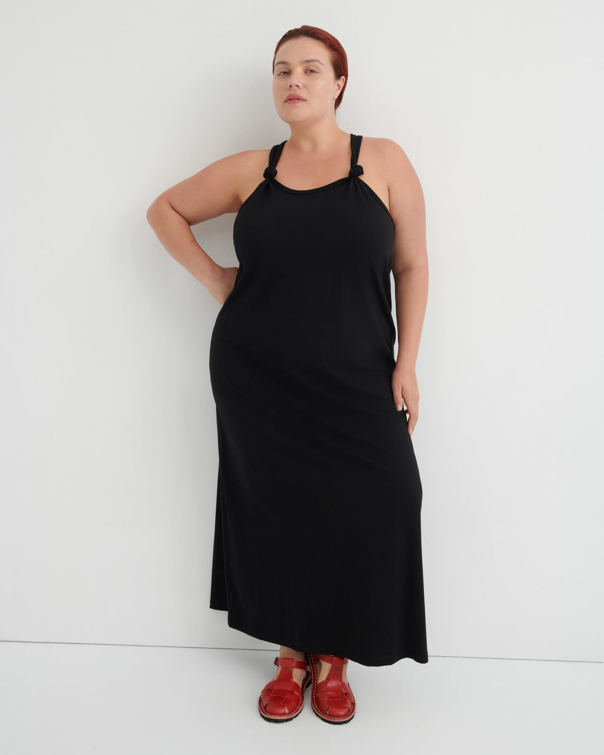 A woman modeling a black halter-neck maxi dress with shoulder cutouts, paired with brown leather sandals, standing against a plain white studio background.