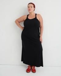 A woman modeling a black halter-neck maxi dress with shoulder cutouts, paired with brown leather sandals, standing against a plain white studio background.