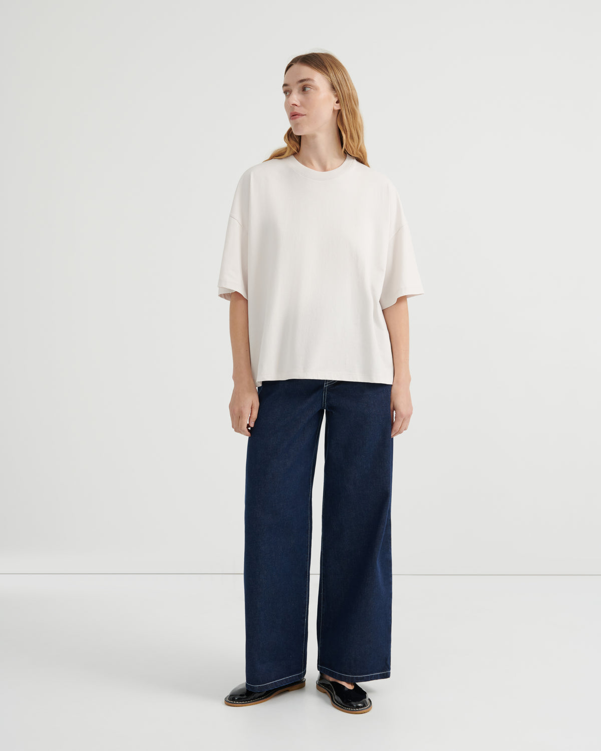 A woman wearing an oversized off-white short-sleeve top, dark blue wide-leg jeans, and black flats.