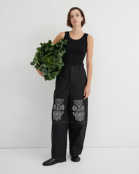 A model stands against a light gray studio background wearing a black sleeveless top and black wide-leg trousers with ornate silver embroidery near the lower legs. She holds a large green leafy vegetable in one hand.