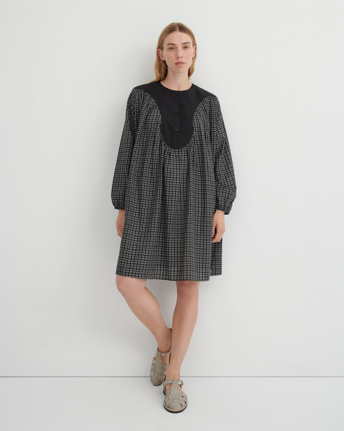 A woman modeling a black and white micro-check dress with a solid black round neckline and buttoned front panel, paired with beige flat sandals, against a plain white studio background.