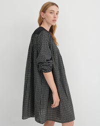 A woman modeling an oversized black and white checkered dress with solid black shoulder yokes, photographed against a neutral light gray studio background.