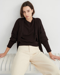 A woman wearing a dark brown ribbed cardigan over a matching dark brown top and cream wide-leg trousers, sitting on a white slatted bench against a light gray wall.