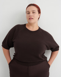 A woman with red hair pulled back wears a dark brown short-sleeve knit top and matching dark brown bottoms, standing against a light gray studio background with her hands in her pockets.